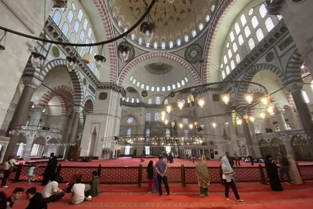 Interior of a grand mosque with high dome, arches, intricate patterns, chandeliers, and people on a red carpeted floor.