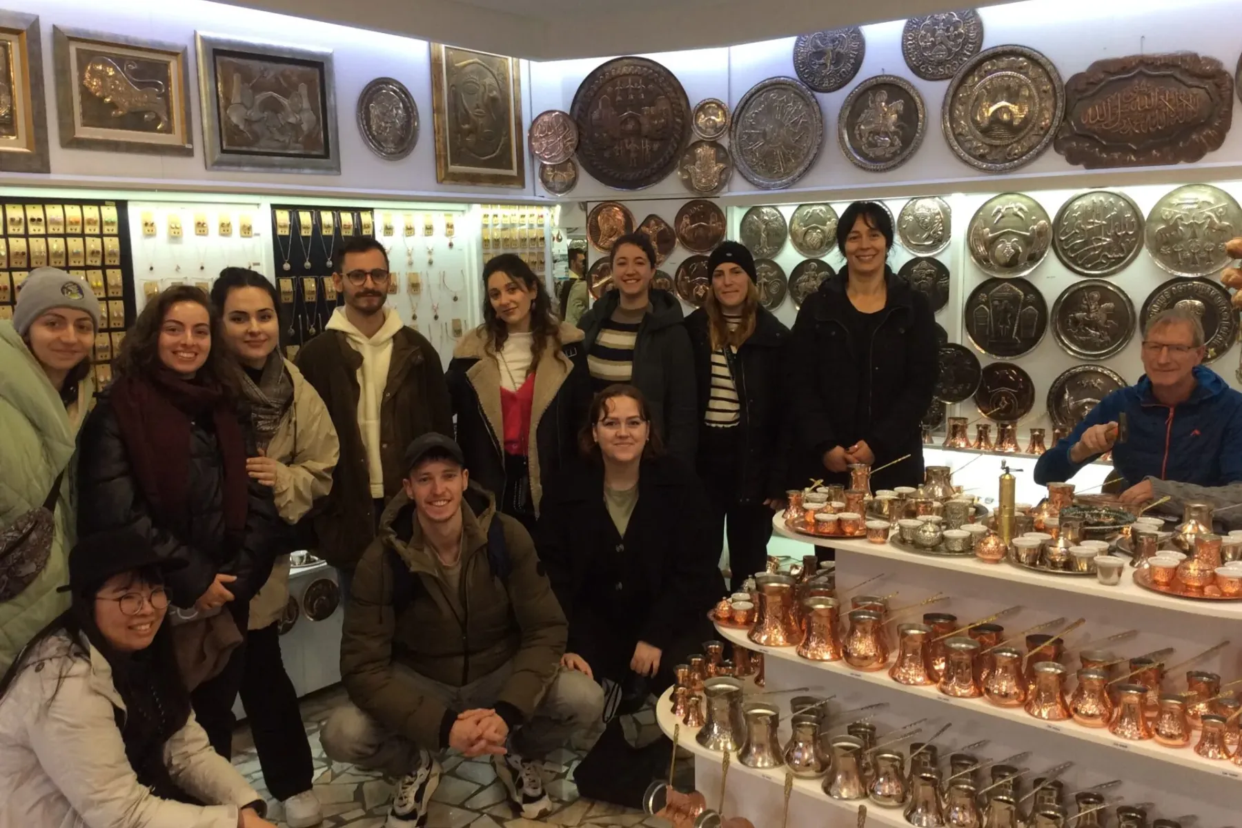 People stand in a shop showcasing numerous copper coffee pots, small cups, and decorative metal plates. An artisan works o...