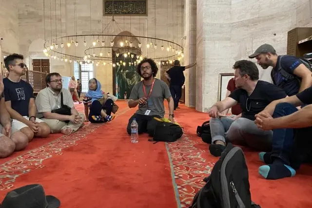 A group of tourists sits on a red carpet inside a large mosque with high ceilings and chandeliers, listening to their guide.
