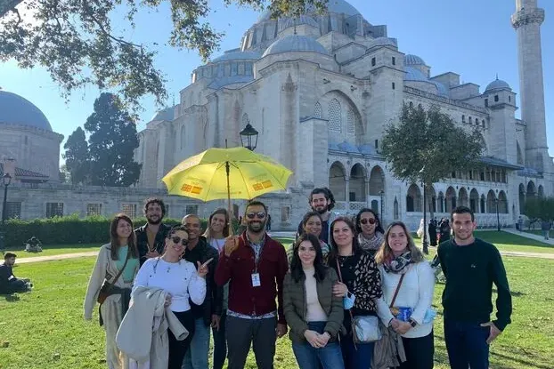 A group of people standing on a grassy lawn in front of the large, domed Süleymaniye Mosque and its minaret in Istanbul.