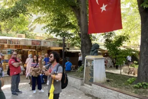 Male tour guide speaking to a group of tourists under green trees. A Turkish flag hangs from a tree; a bust statue is nearby.