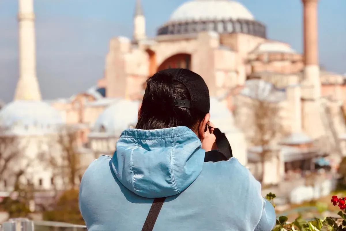 Man in blue hoodie and black cap views Hagia Sophia and its minarets under a clear sky from a terrace.