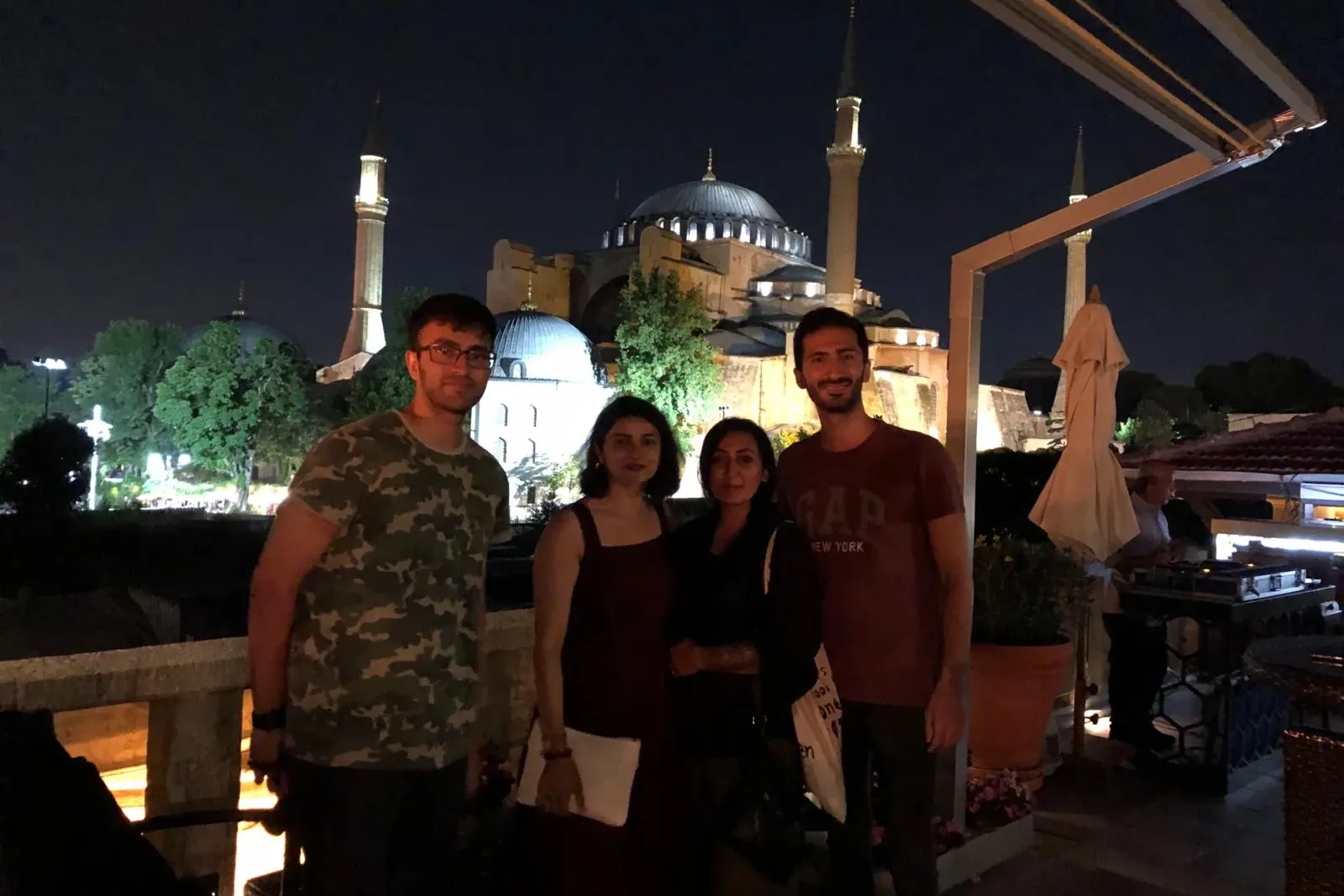 Four people standing on a balcony at night, with the illuminated Hagia Sophia and its minarets in the background.