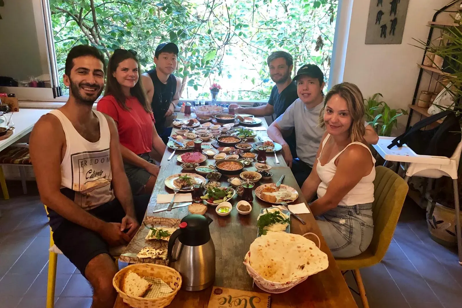 Six people sit at a long table laden with a variety of small dishes, flatbread, and tea, with green foliage visible outside.