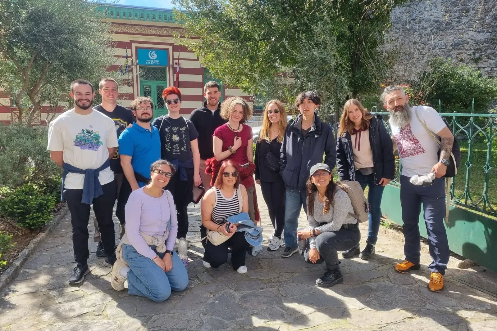 A diverse group of people, including men and women, poses for a photo outdoors on a stone path in Mostar, with a striped b...