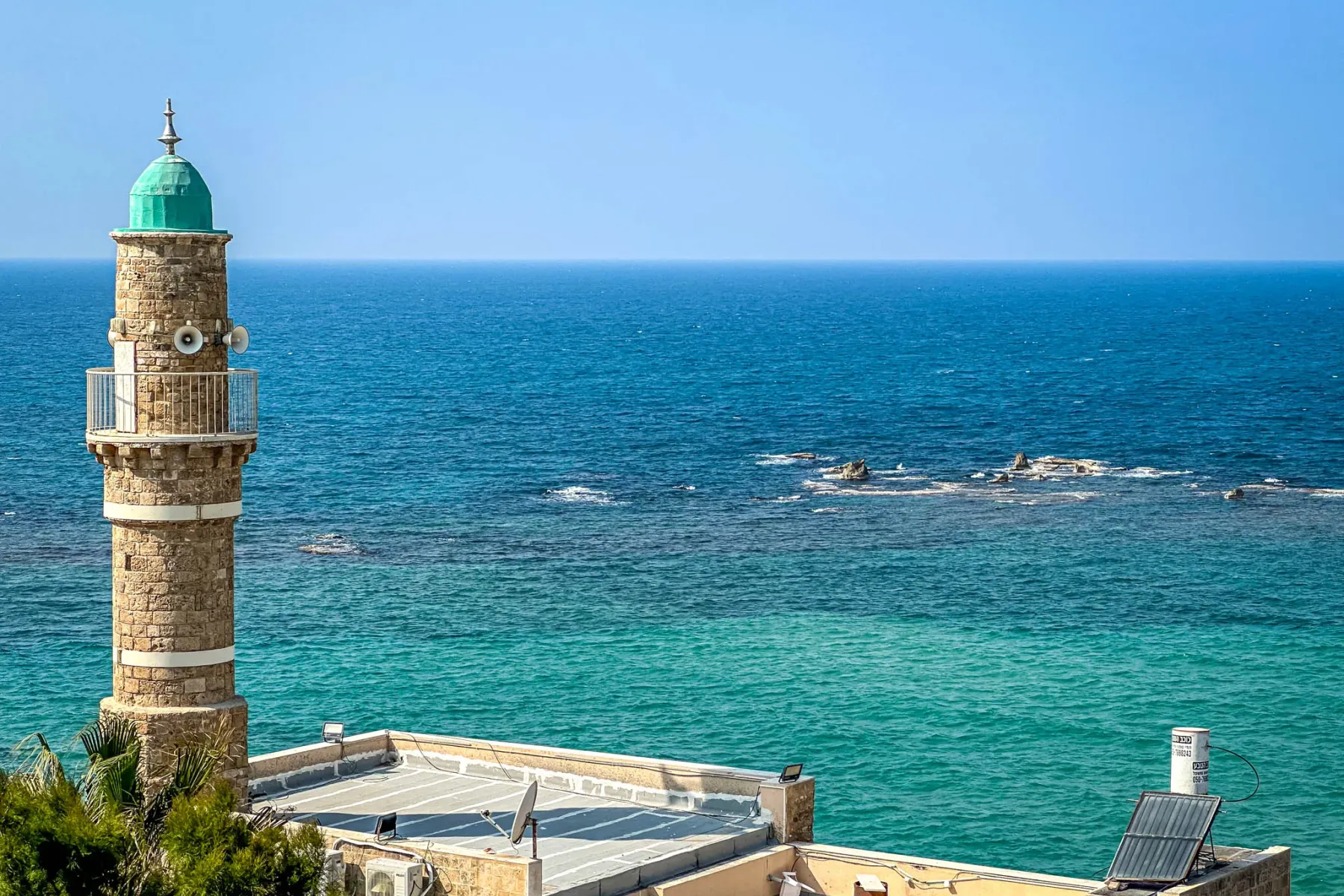 Stone minaret with a green dome and balcony overlooking the turquoise Mediterranean Sea, with rooftops and green foliage.