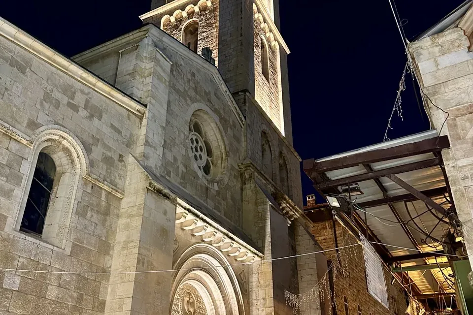 Illuminated stone church with a tall bell tower, arched entrance, and rose window at night in Jerusalem. Adjoining old cit...