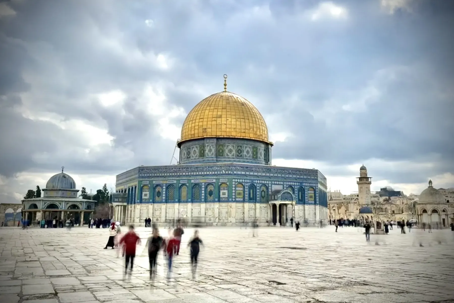 The Dome of the Rock with its golden dome and blue tiles under a cloudy sky, people walking on a stone plaza.