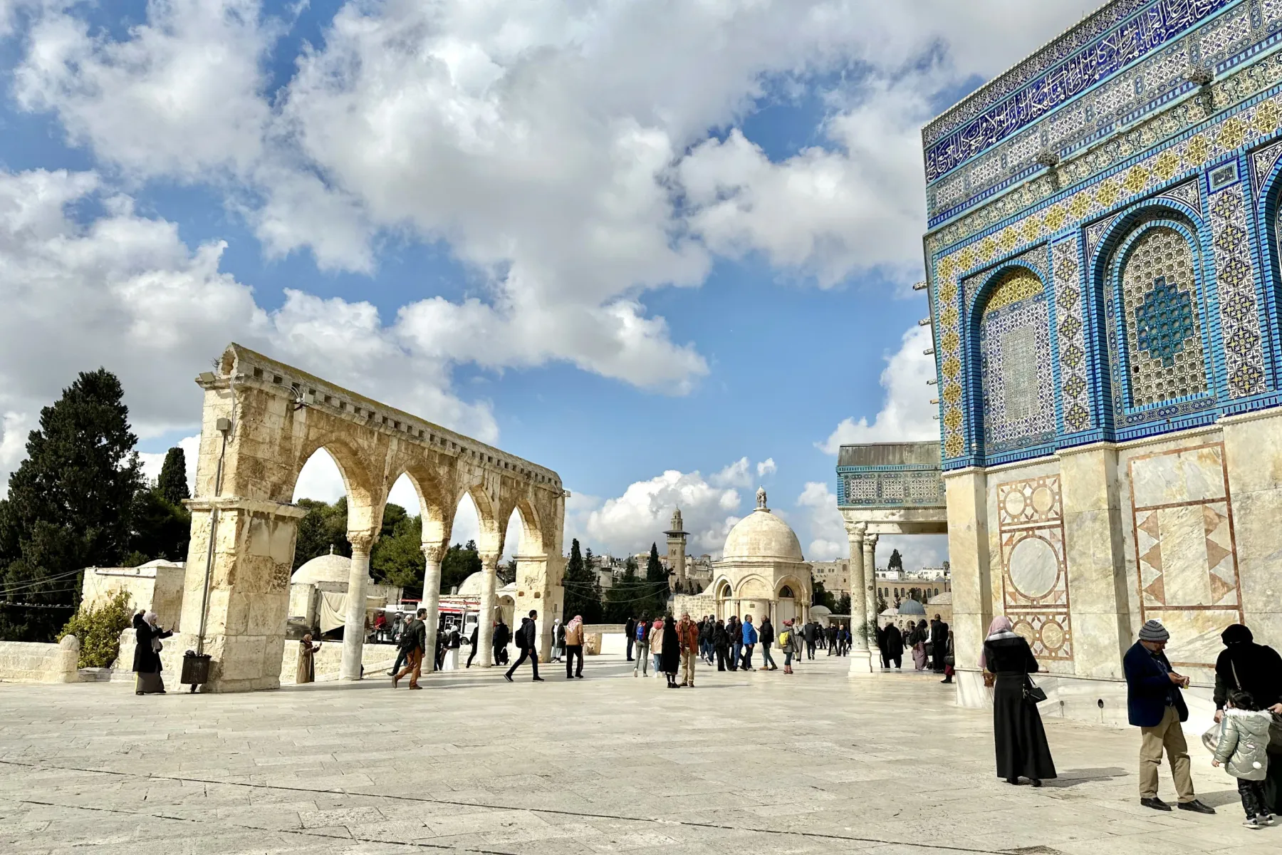 People walk on the wide plaza of Temple Mount, framed by stone arches on the left and the ornately tiled facade of the Dom...