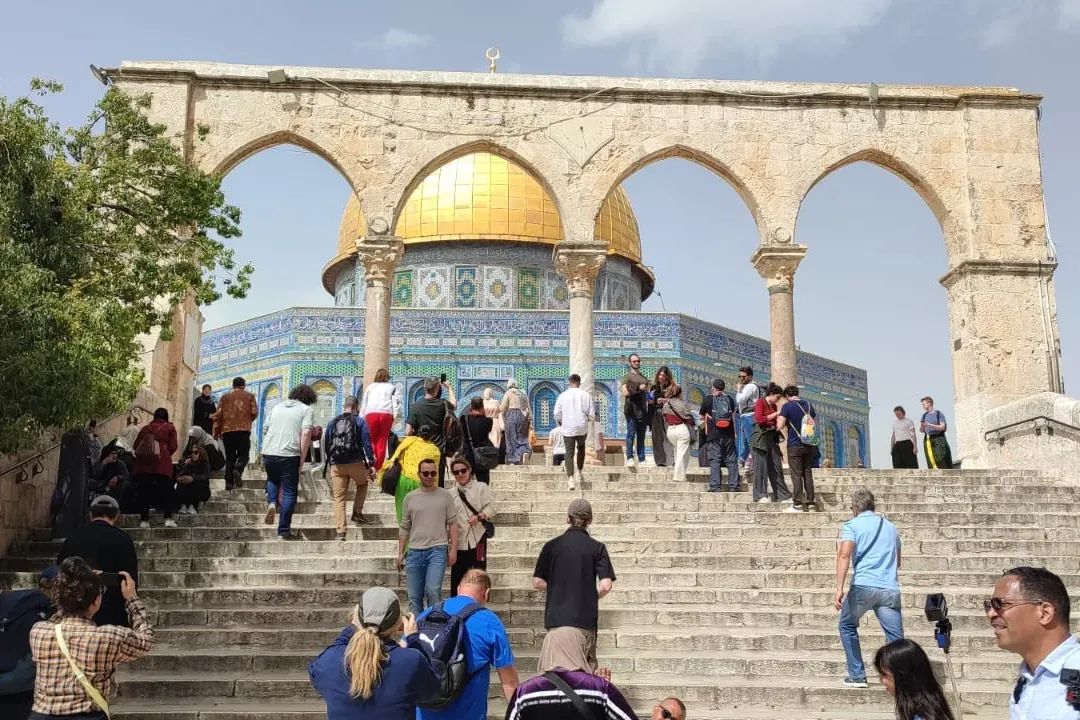 Tourists ascend stone stairs towards the Dome of the Rock with its golden dome and blue tiled facade, framed by ancient st...