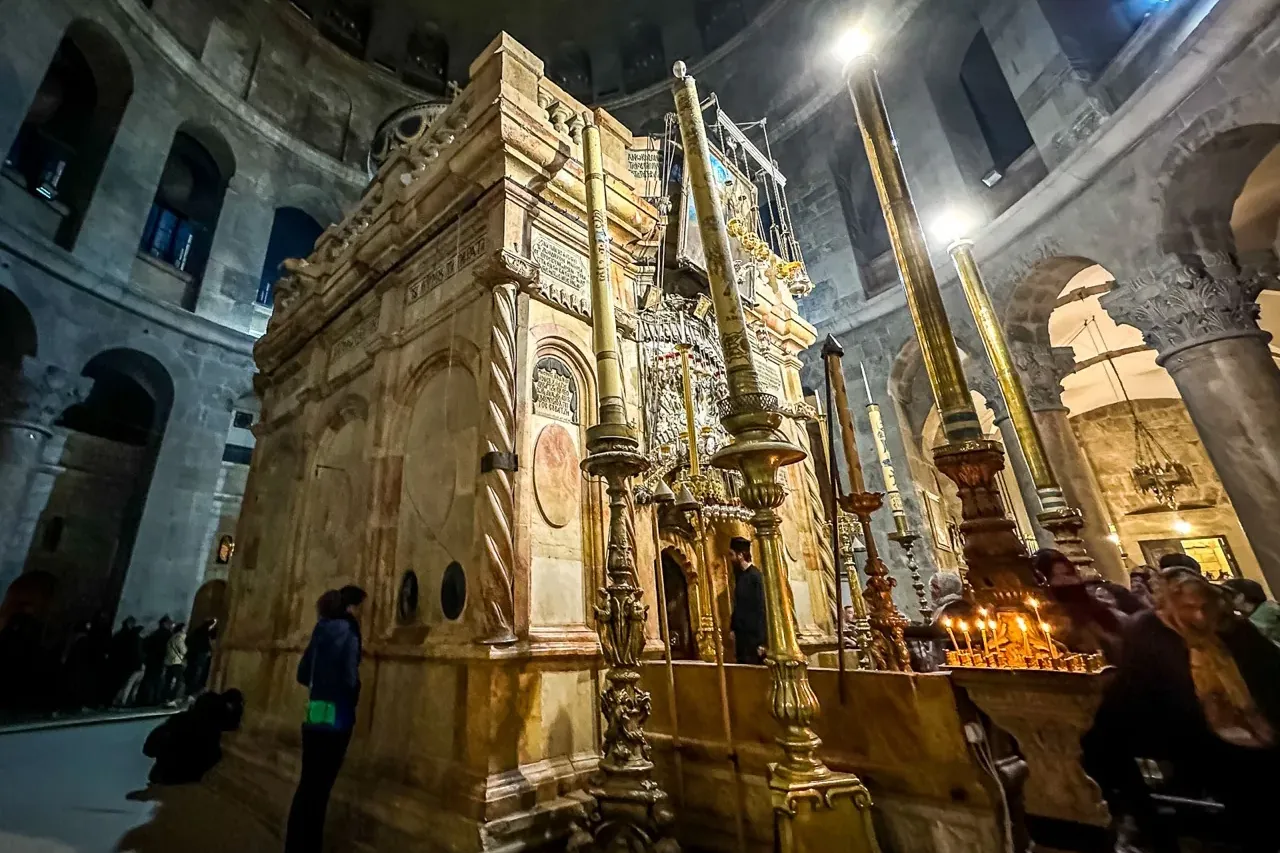 The stone Edicule inside the Church of the Holy Sepulchre, illuminated by tall candles and artificial lights, with people ...