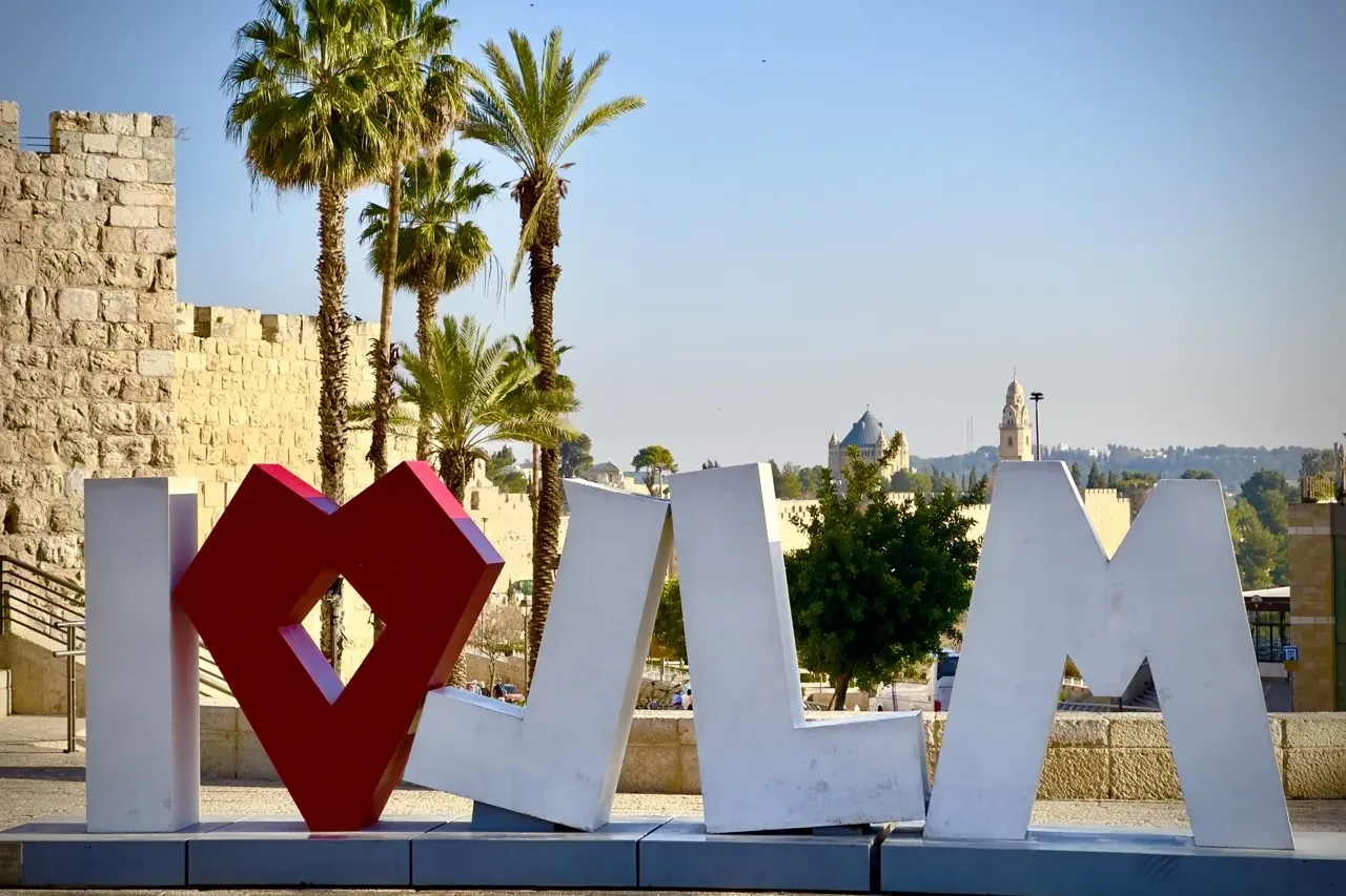 Large white 'I' and 'JLM' letters with a red heart symbol in a public space. Stone wall and palm trees are behind, distant...