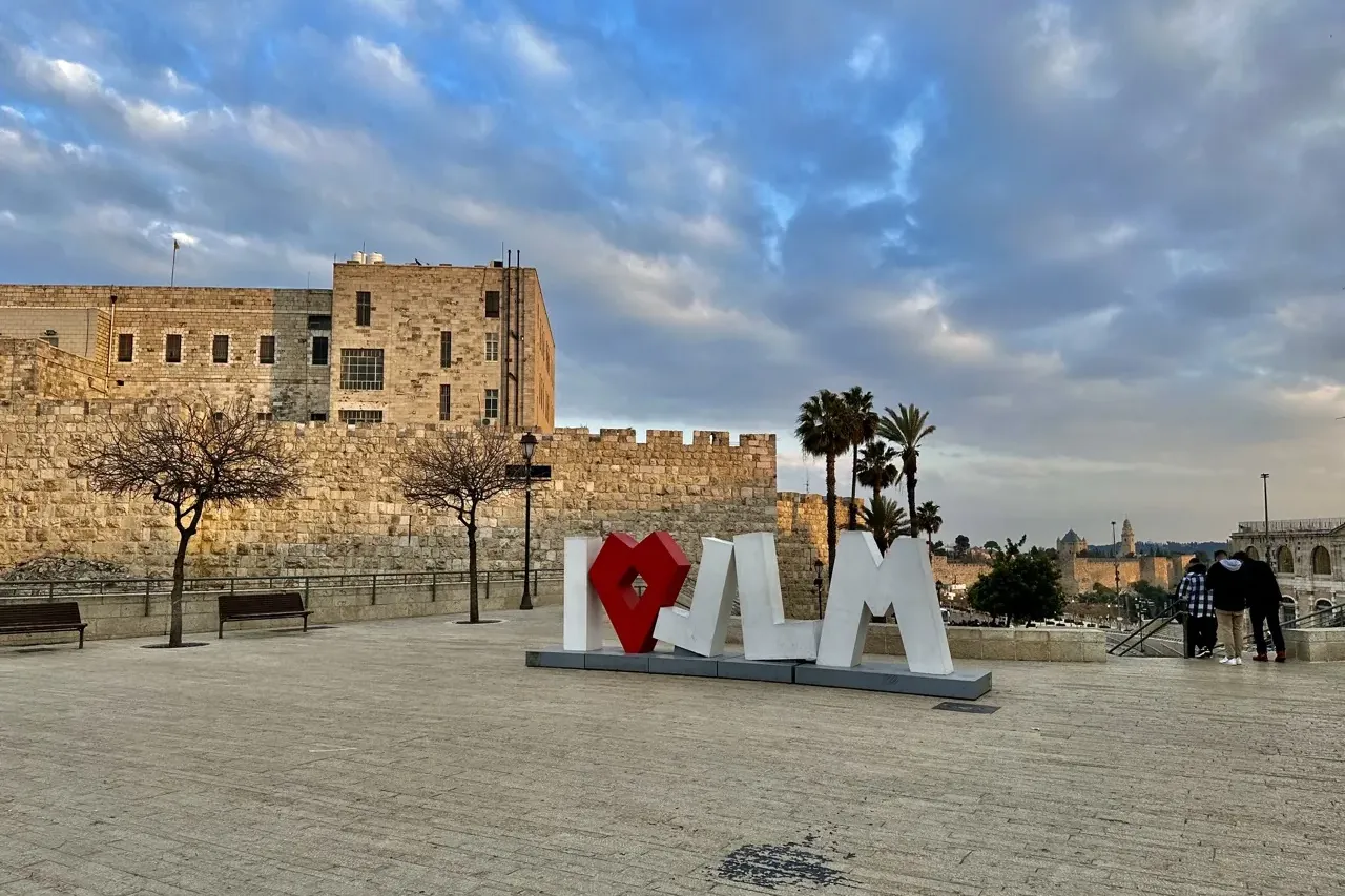 Large white I love JLM sign with a red heart, in a paved square. Behind it are ancient stone walls, buildings, and bare tr...