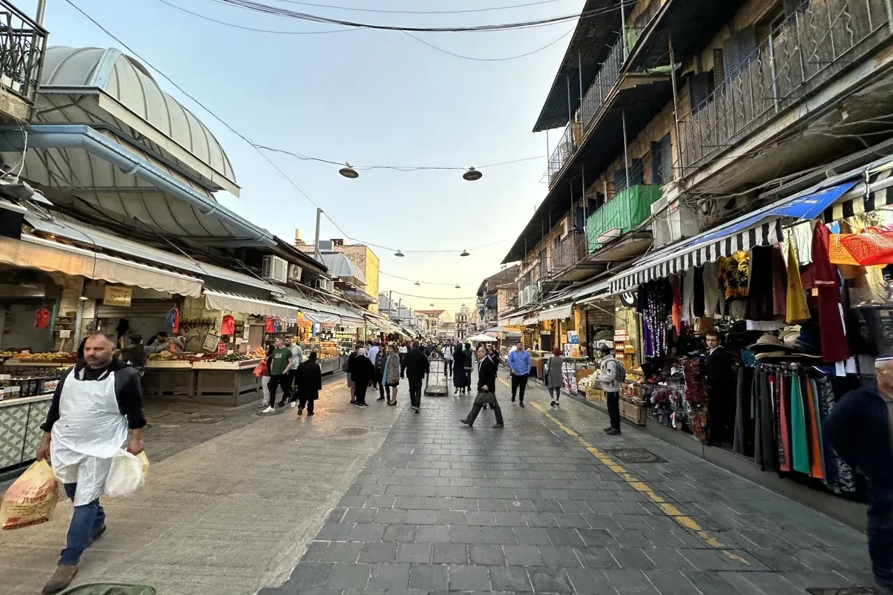 Bustling outdoor market street in Jerusalem with stalls selling produce and goods, people walking, and multi-story buildin...