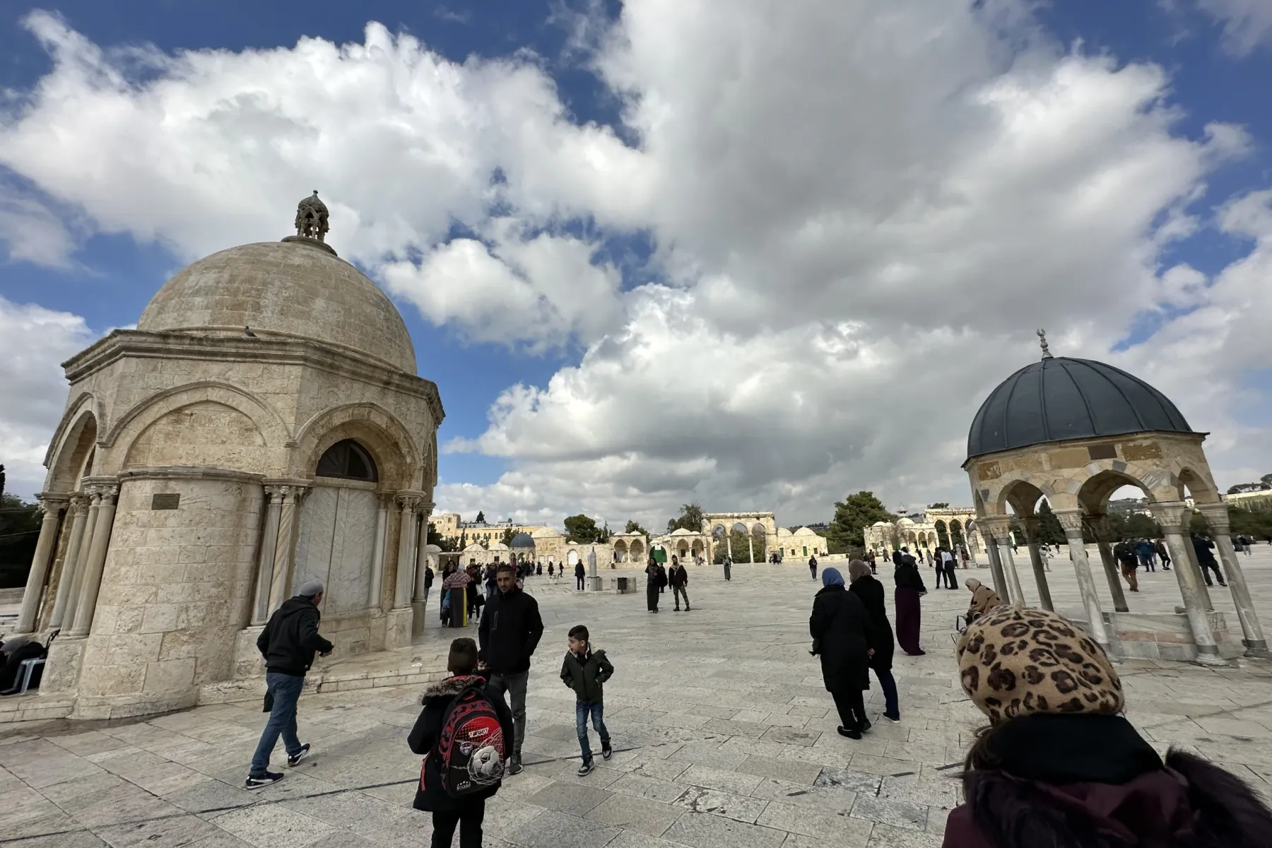 People walk on a large stone plaza with two domed stone structures and distant arched buildings under a cloudy blue sky.