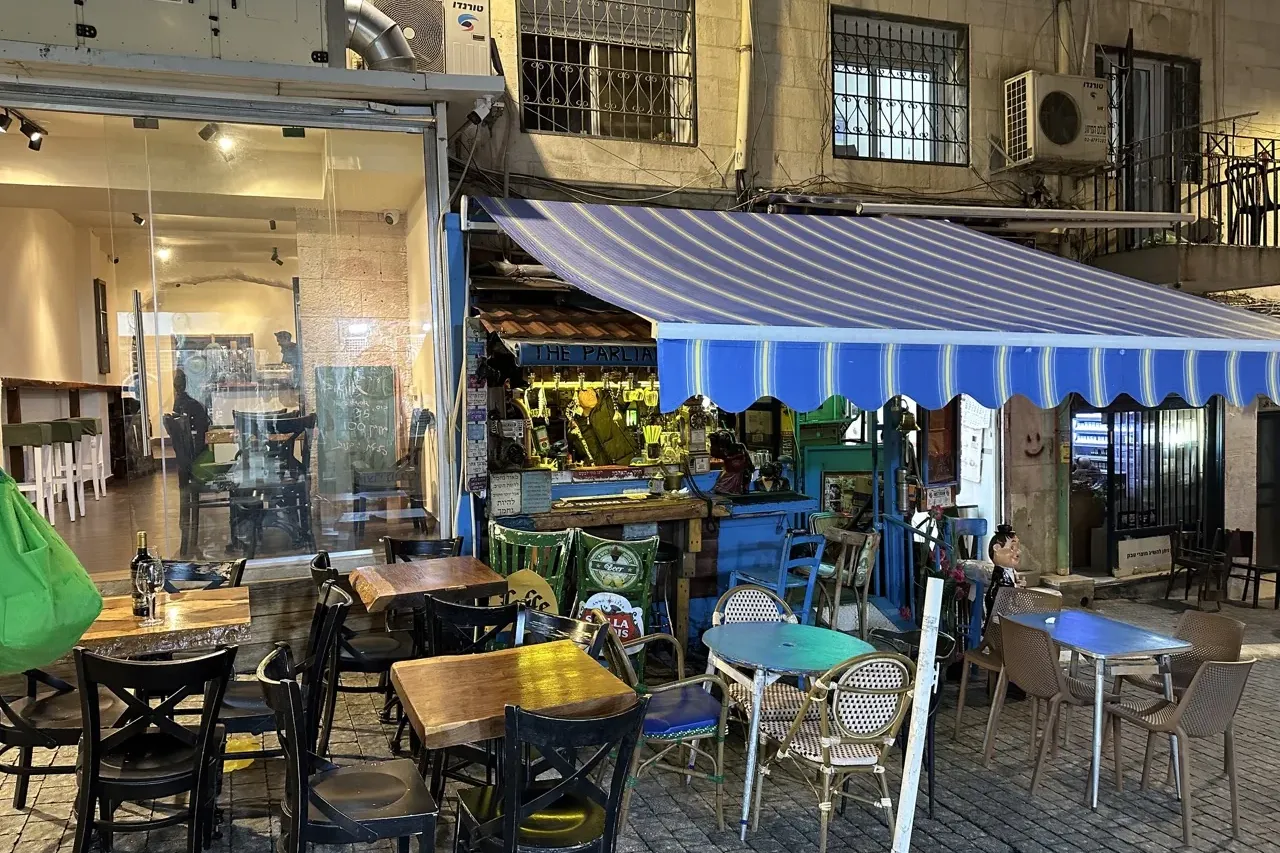 Outdoor bar or cafe with various tables and chairs under a blue and white striped awning at night. A building facade is vi...