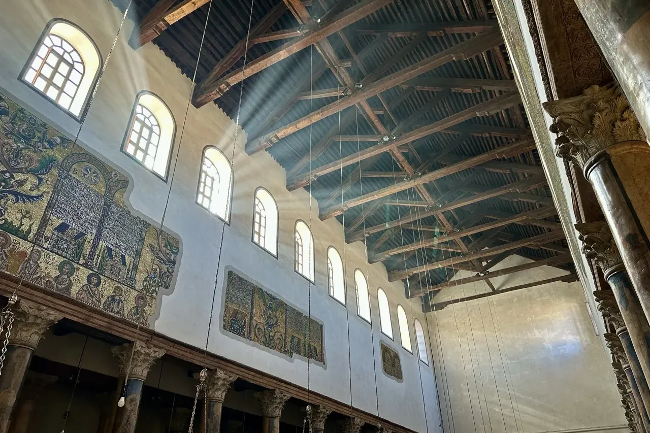 Interior of a church with ancient golden mosaics, arched windows, and a dark wooden beamed ceiling illuminated by sun rays.