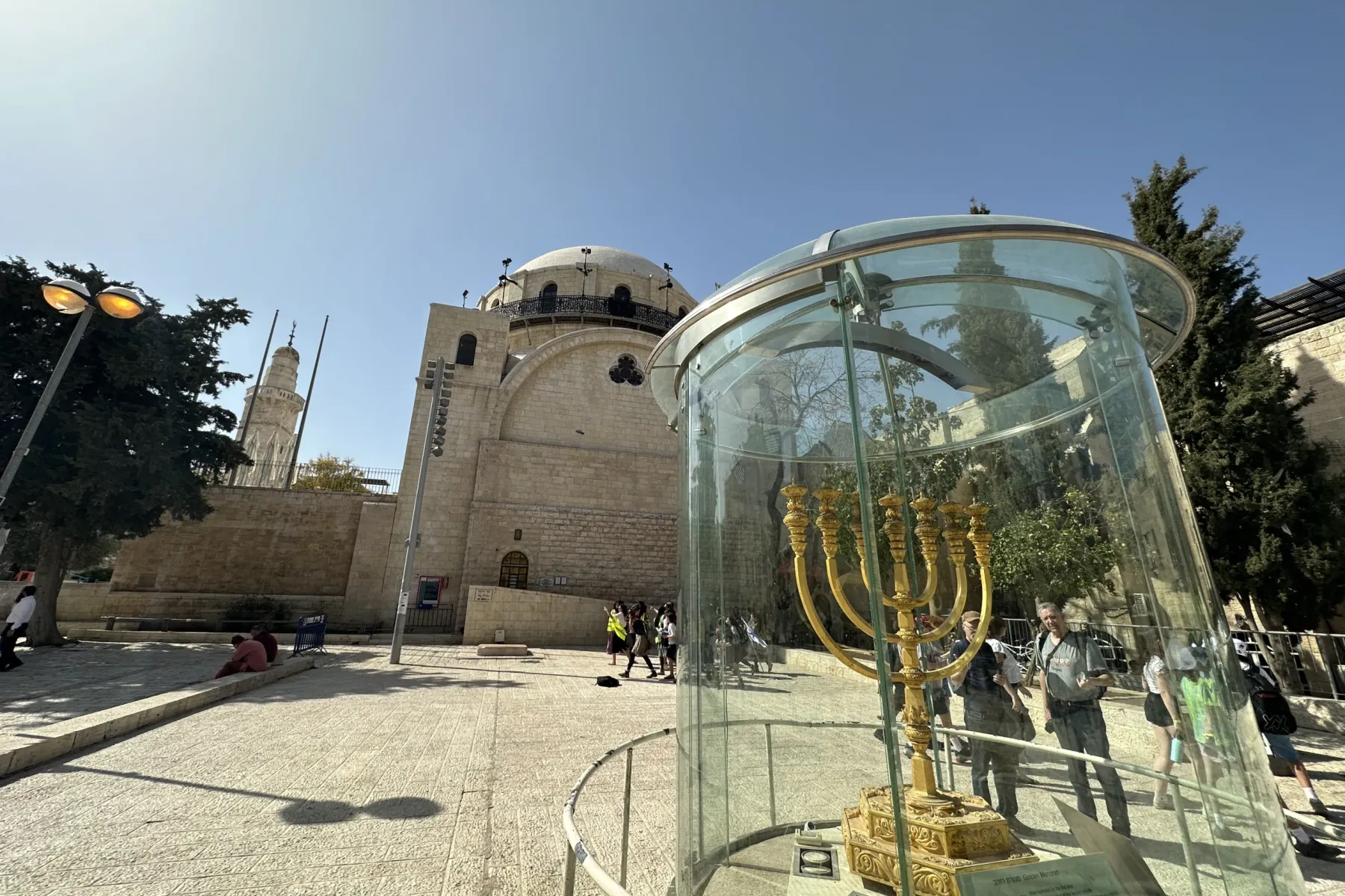Golden menorah in a glass enclosure on a paved plaza, with a stone building and minaret under a clear sky.