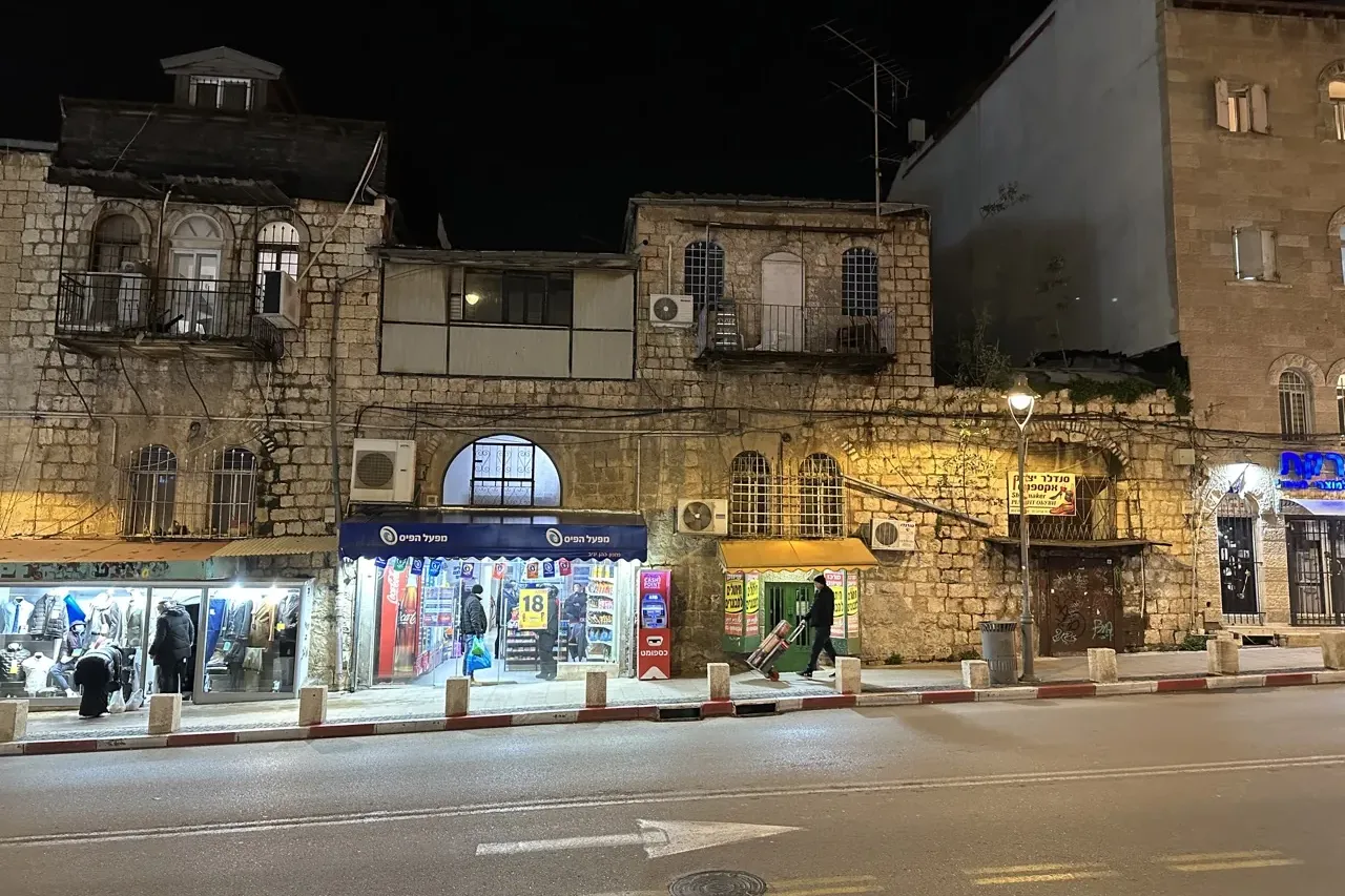 Night view of a street in Jerusalem with historic stone buildings, brightly lit ground-floor shops, and a person walking o...