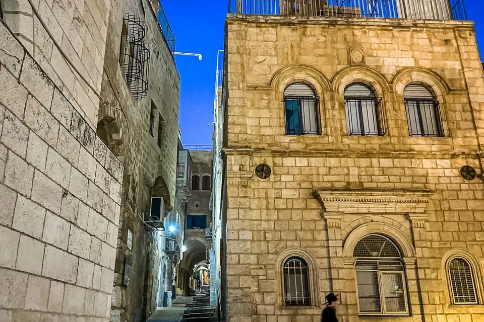Stone buildings flank a narrow, stepped alleyway in Jerusalem at night. A person walks down steps on the right, under a da...