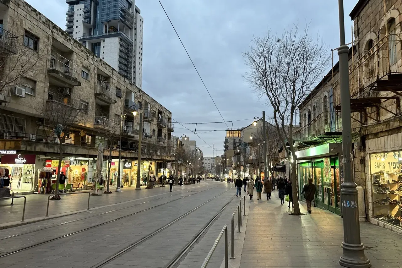 A city street in Jerusalem at dusk, with tram tracks, stone buildings housing shops, pedestrians on sidewalks, and a moder...