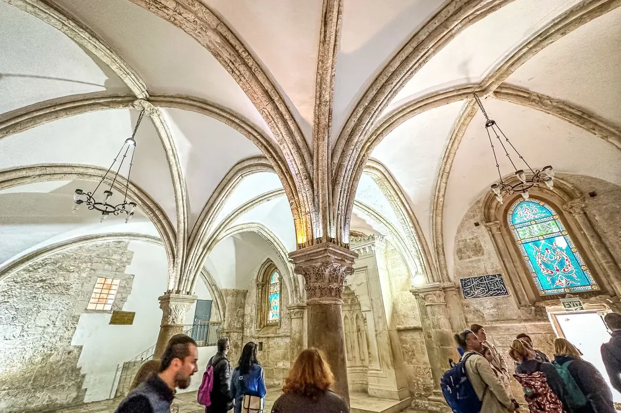 Tour group in a historic vaulted hall with stone walls, ribbed ceiling, a blue stained glass window, and Arabic script.