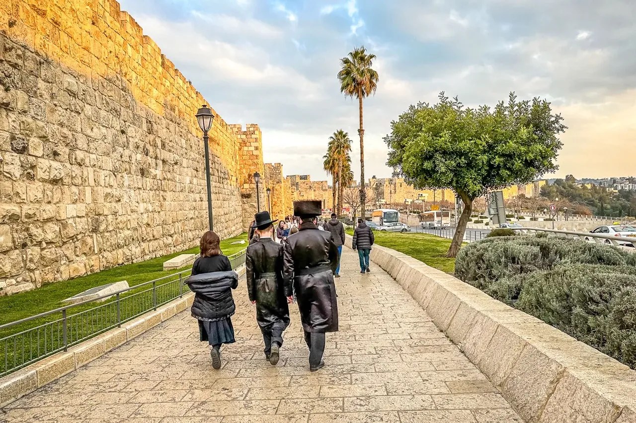 People walk along a paved path next to Jerusalem's Old City walls, with palm trees and a cloudy sky above.