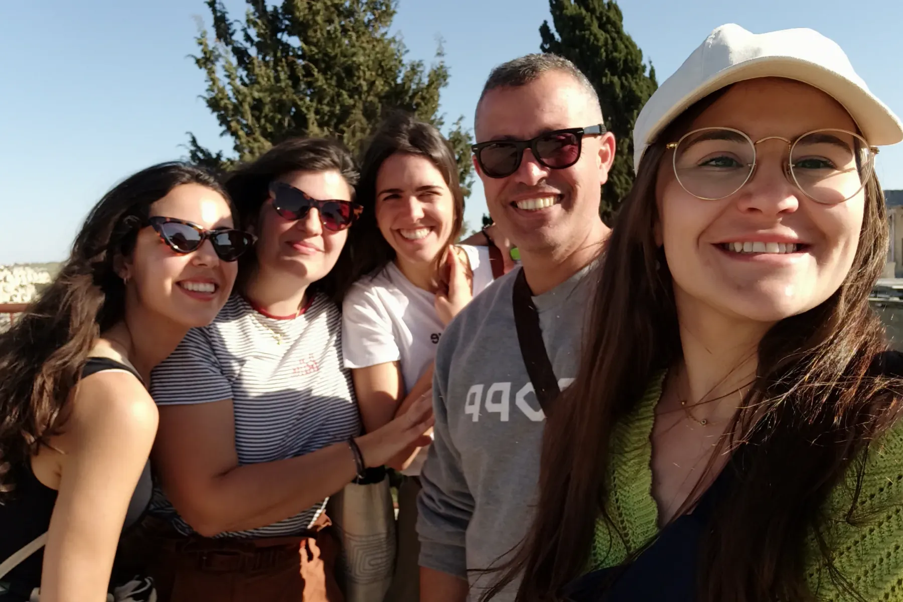 Five smiling people, four women and one man, posing for a selfie outdoors with a distant town and trees under a clear blue...