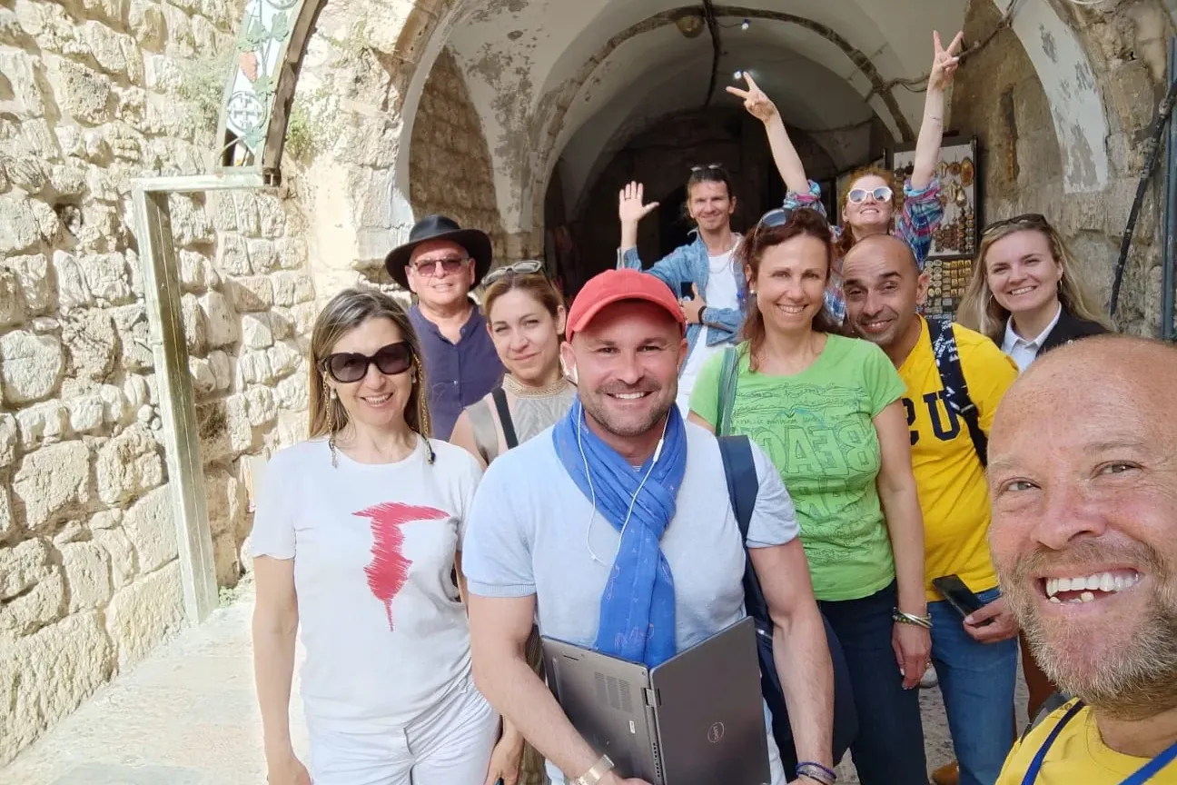 Group of smiling tourists and a guide taking a selfie in a narrow stone alleyway with arches in Jerusalem Old City.