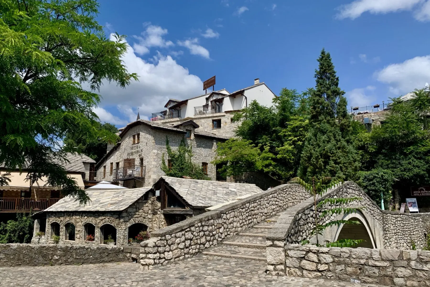 Stone bridge with steps leading to historic stone buildings and a hotel under a blue sky with white clouds.