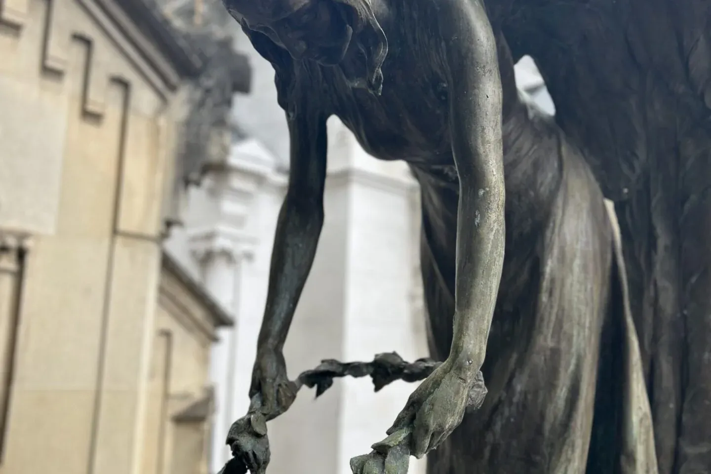 Close-up of a weathered bronze sculpture of an angel with wings, bowing while placing a leafy wreath over a supine figure.