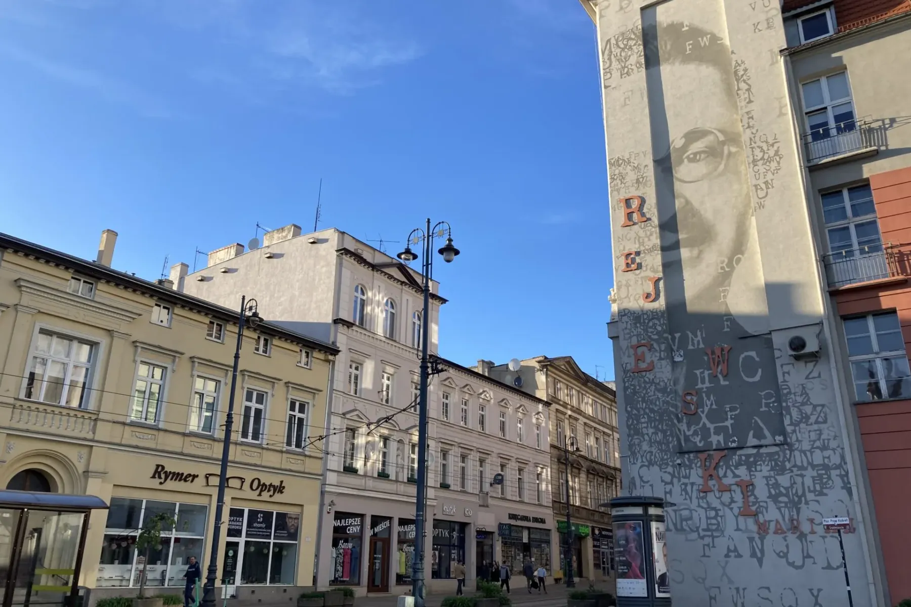 Street scene in Bydgoszcz with historic buildings, a prominent mural of Mikołaj Rej on a building, shops, and a tram stop.