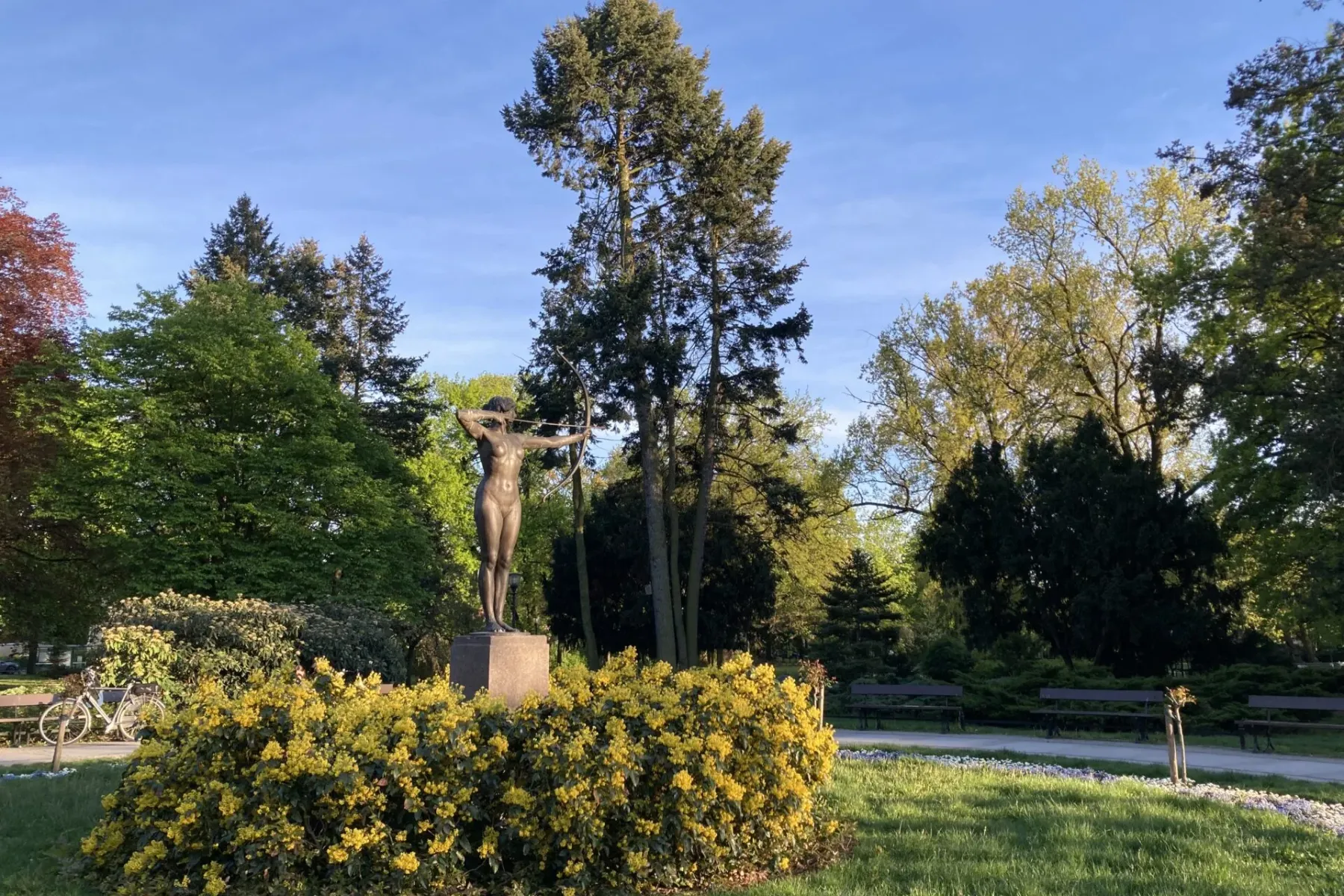 Bronze statue of an archeress in a park, surrounded by yellow flowering bushes, green trees, and grass under a blue sky.