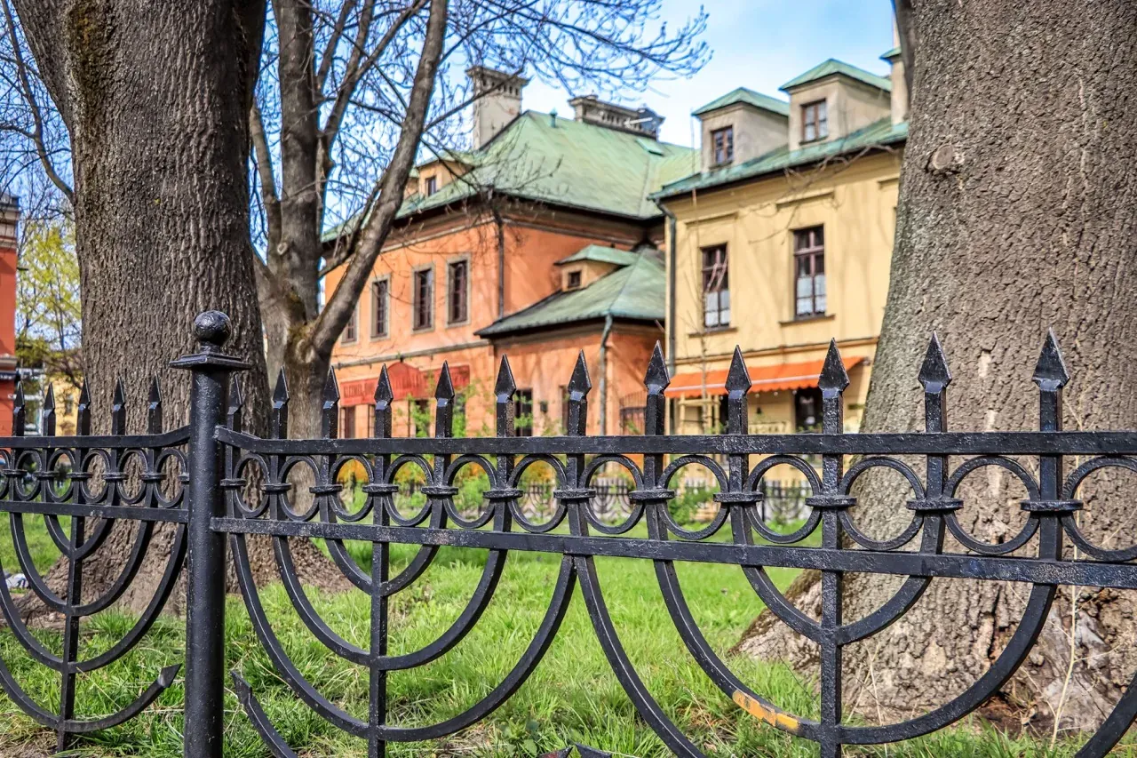 Black wrought-iron fence with stylized menorah-like design in foreground, green grass, tree trunks, and historic buildings...