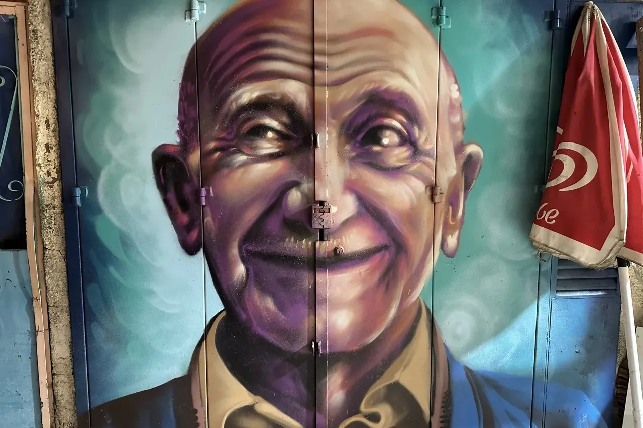 Colorful mural of a smiling bald elderly man on blue metal doors, with a folded red umbrella on the right.