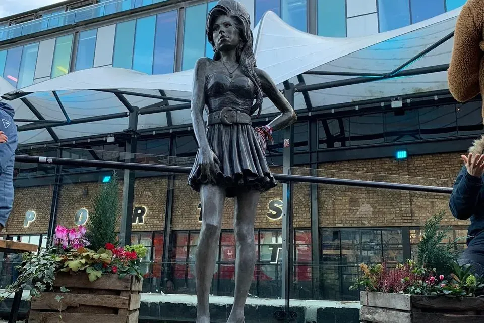 Bronze statue of Amy Winehouse with her hand on her hip, standing on paved ground in front of modern buildings under a clo...