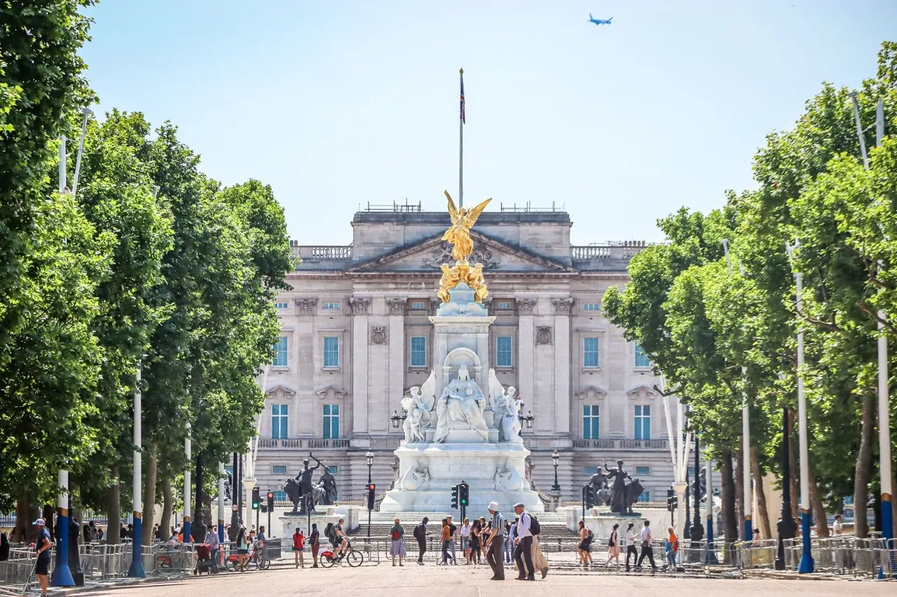 Buckingham Palace and the Victoria Memorial with people walking around on a sunny day in London. An airplane flies overhead.