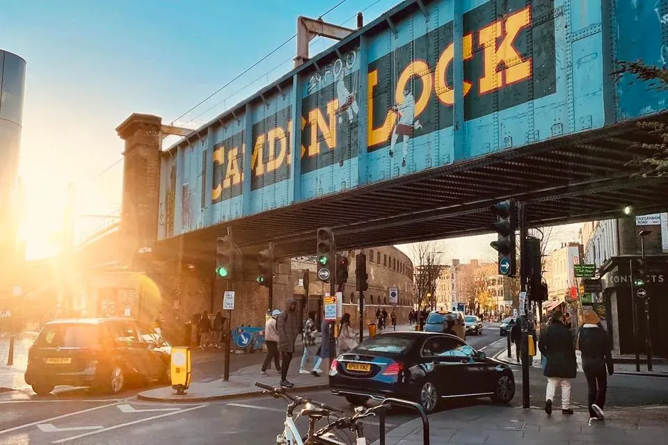 Blue railway bridge with "CAMDEN LOCK" painted on it, spanning a street with cars, pedestrians, and parked bicycles at sun...