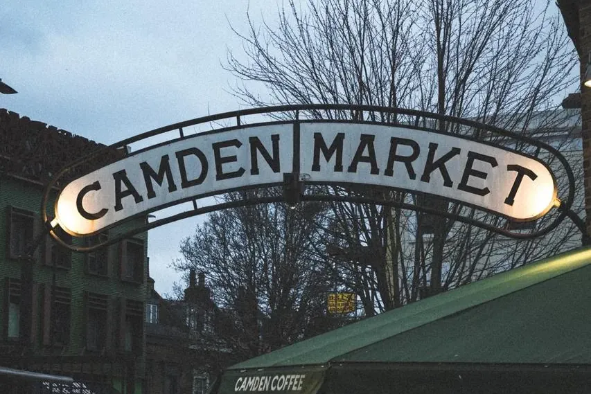 Illuminated archway sign for Camden Market at dusk, with bare trees, buildings, and people in the foreground.
