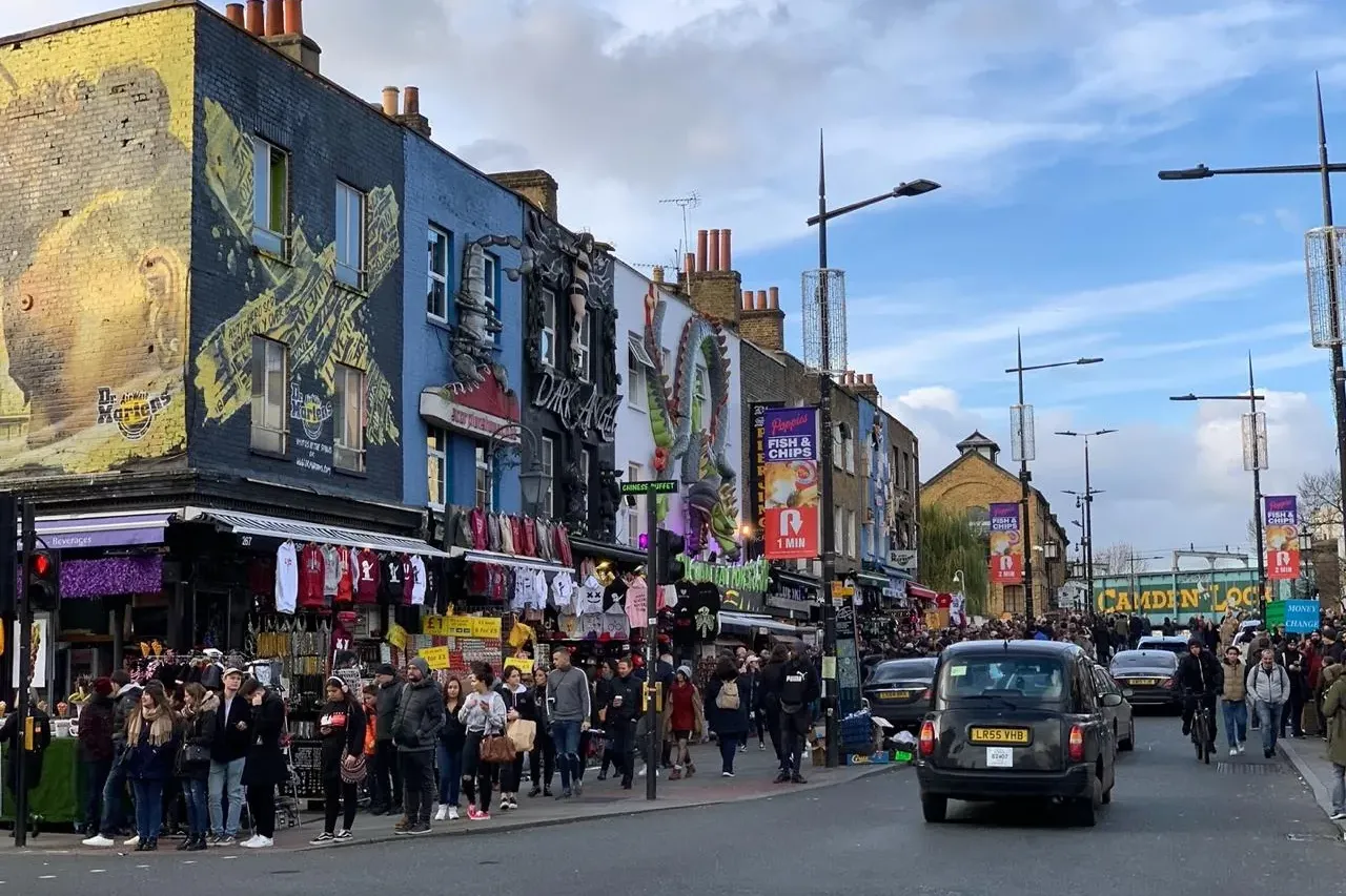Busy street in London's Camden Town with colorful shop facades, many pedestrians, a black taxi, and market stalls.