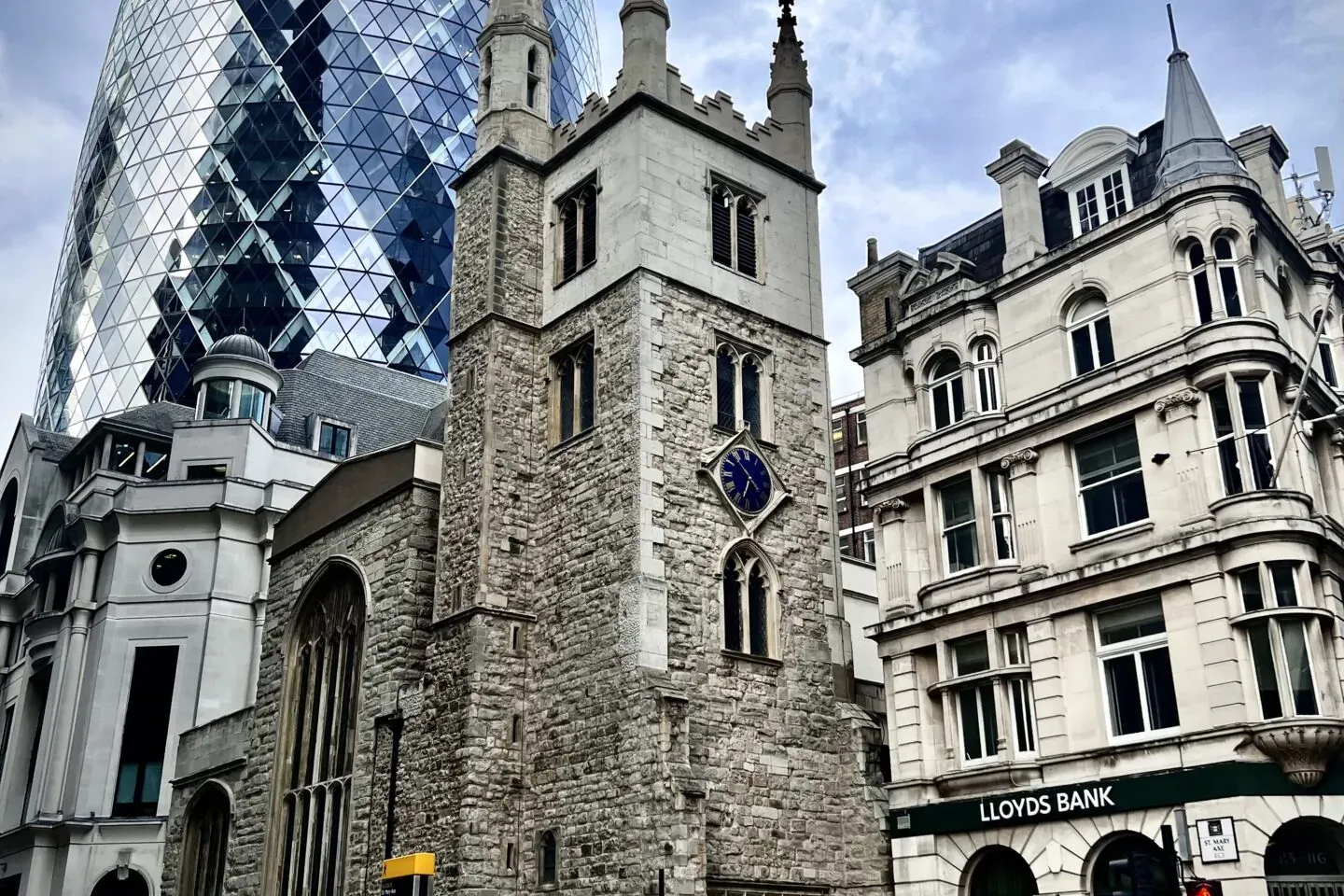 The Gherkin skyscraper, a historic stone church tower with a blue clock, and a Lloyds Bank building on a busy London street.