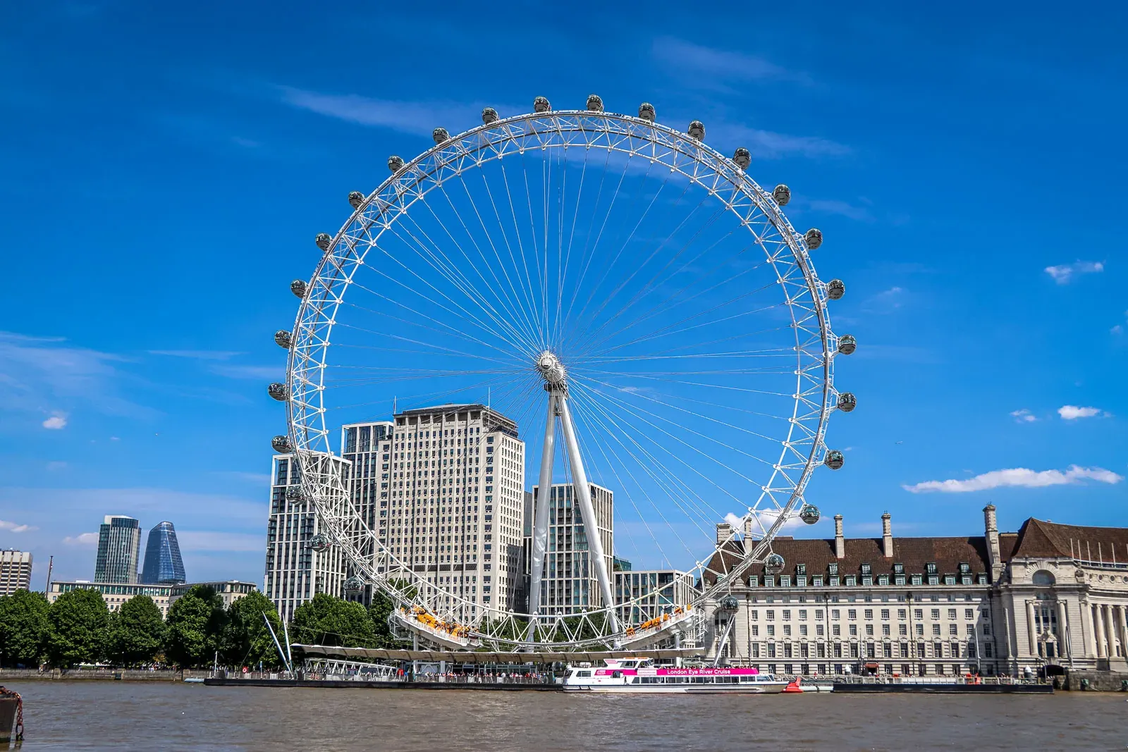 The London Eye Ferris wheel on the River Thames bank, with modern and historic buildings under a blue sky.