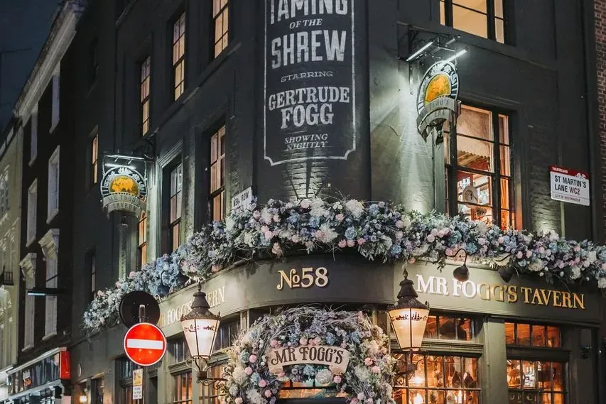 Exterior of Mr Fogg's Tavern in London at night, decorated with blue and pink floral garlands around the entrance and wind...