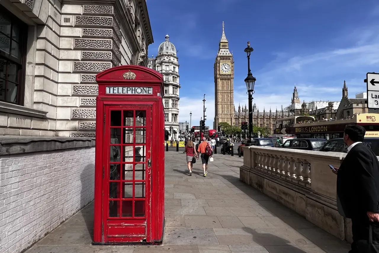 A classic red telephone booth on a London sidewalk with Big Ben and the Houses of Parliament in the background under a blu...