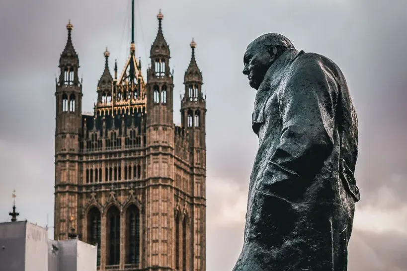 Bronze statue of Winston Churchill with the Houses of Parliament and a Union Jack flag visible in the background under an ...