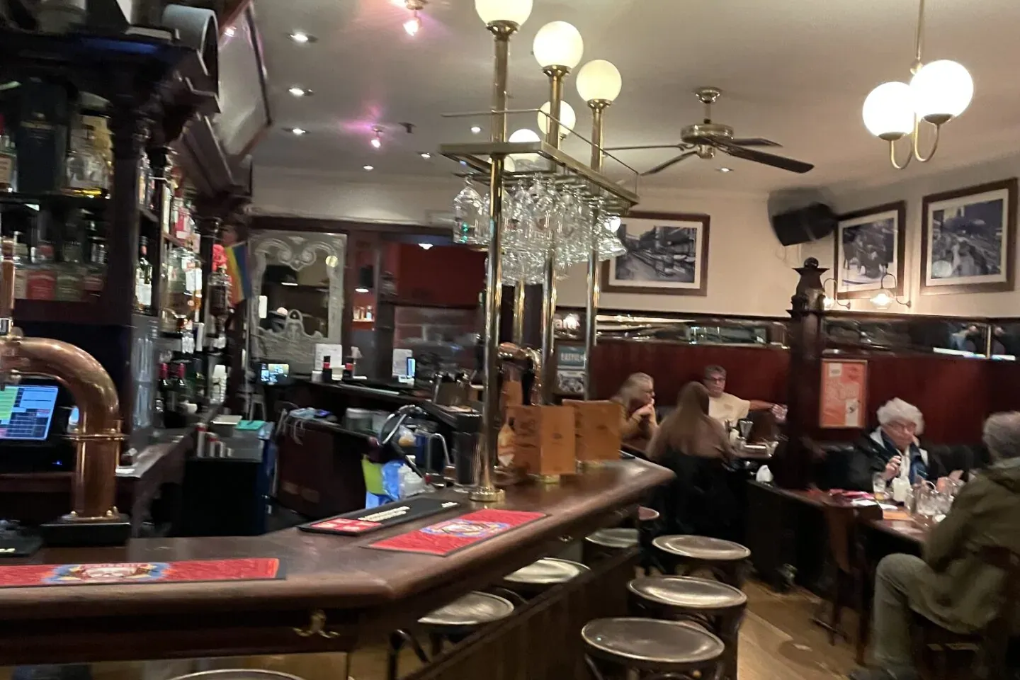 Interior of a traditional pub with a long wooden bar, several empty bar stools, and people seated at tables.