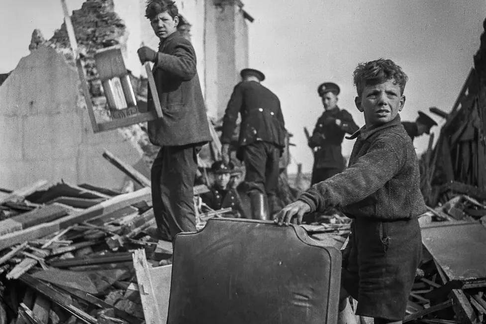 Black and white photo of two boys amidst extensive building rubble and debris. Collapsed walls stand in the background, wi...