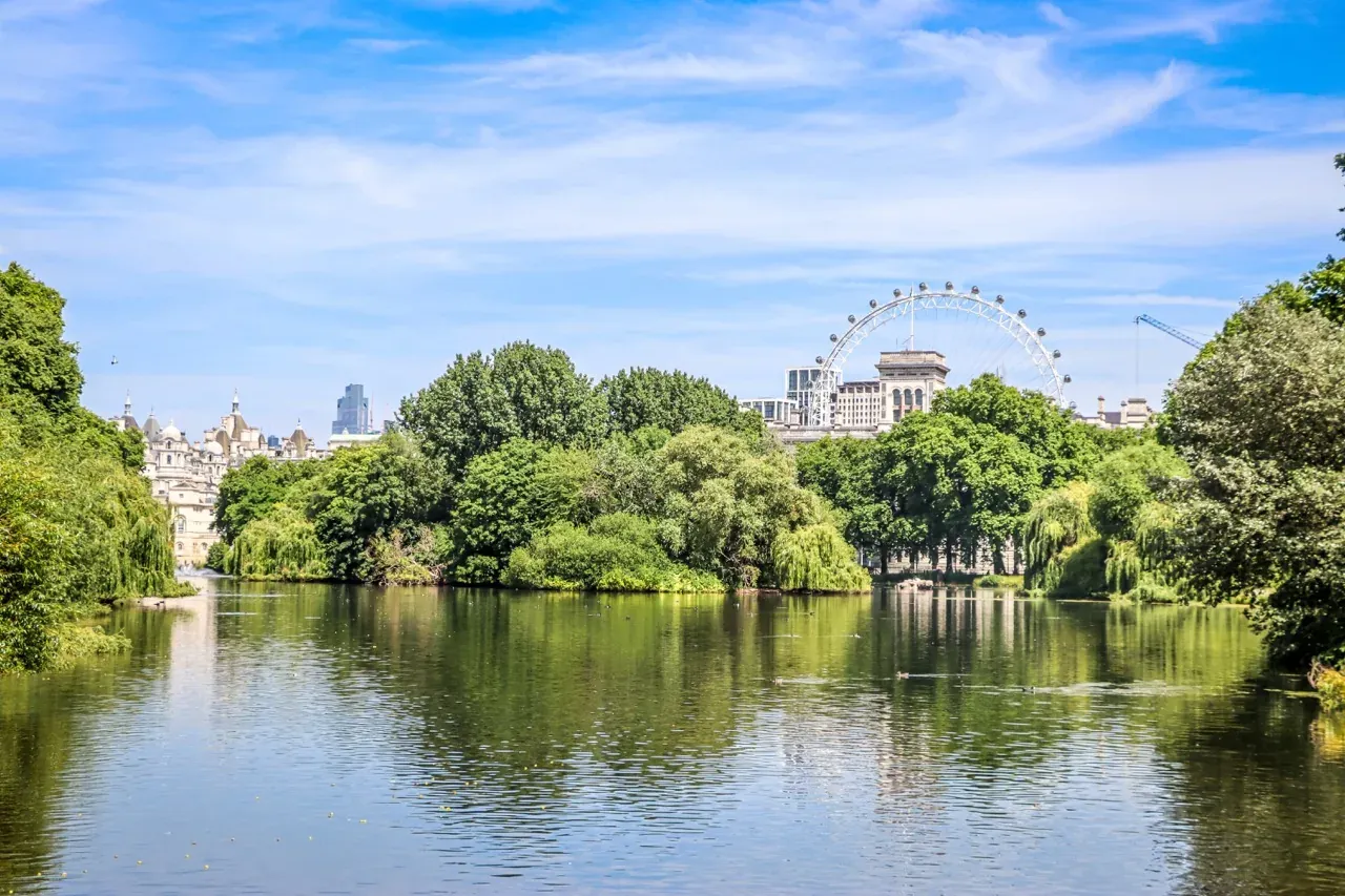A wide park lake with green trees on its banks. The London Eye and buildings of the city skyline are visible under a blue ...