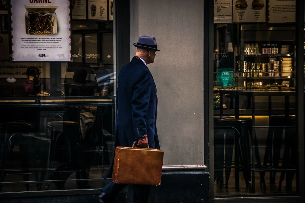 Man in a dark coat and hat walks past a storefront, carrying a brown leather briefcase.