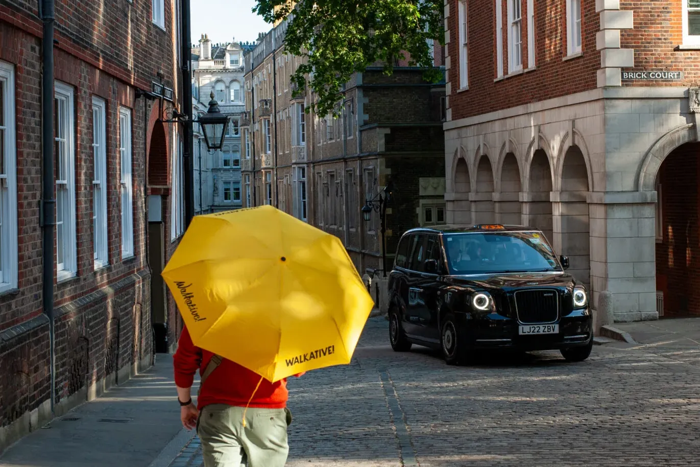 Person with yellow umbrella on a cobblestone street in London's Brick Court, with brick buildings and a black taxi.
